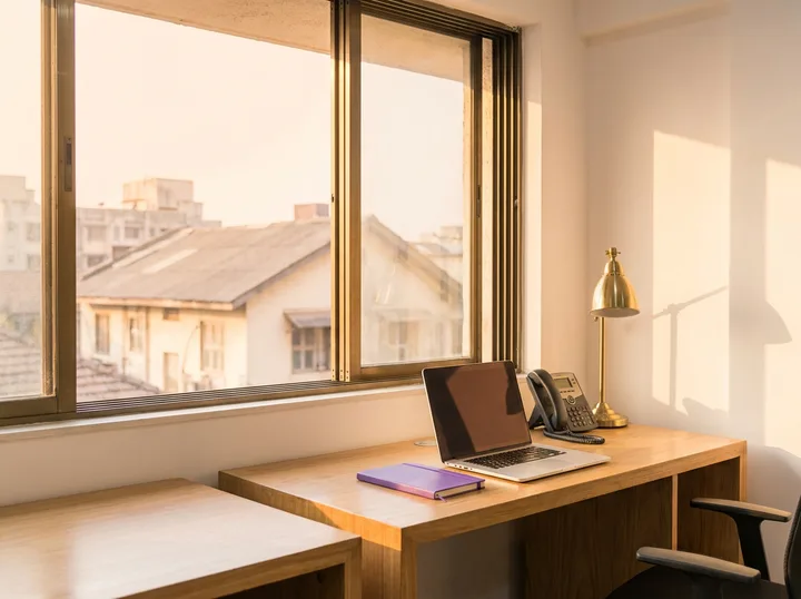 A sunlit desk by a window with a laptop, notebook and brass lamp, looking out over low rooftops