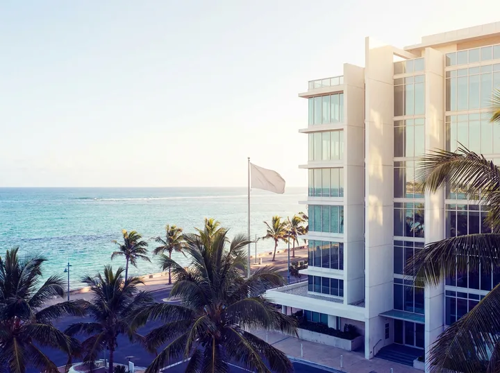 A white office building on a palm-lined coastal road above a turquoise sea